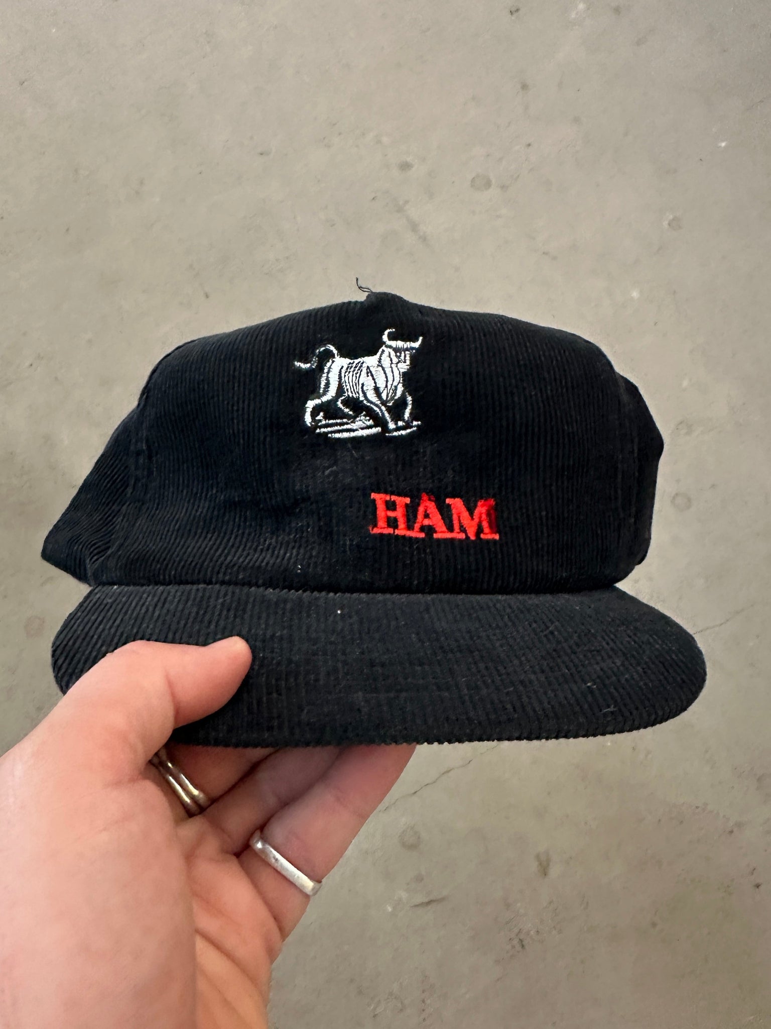 Black cap with a bull logo and 'HAM' text held by a hand on a concrete surface