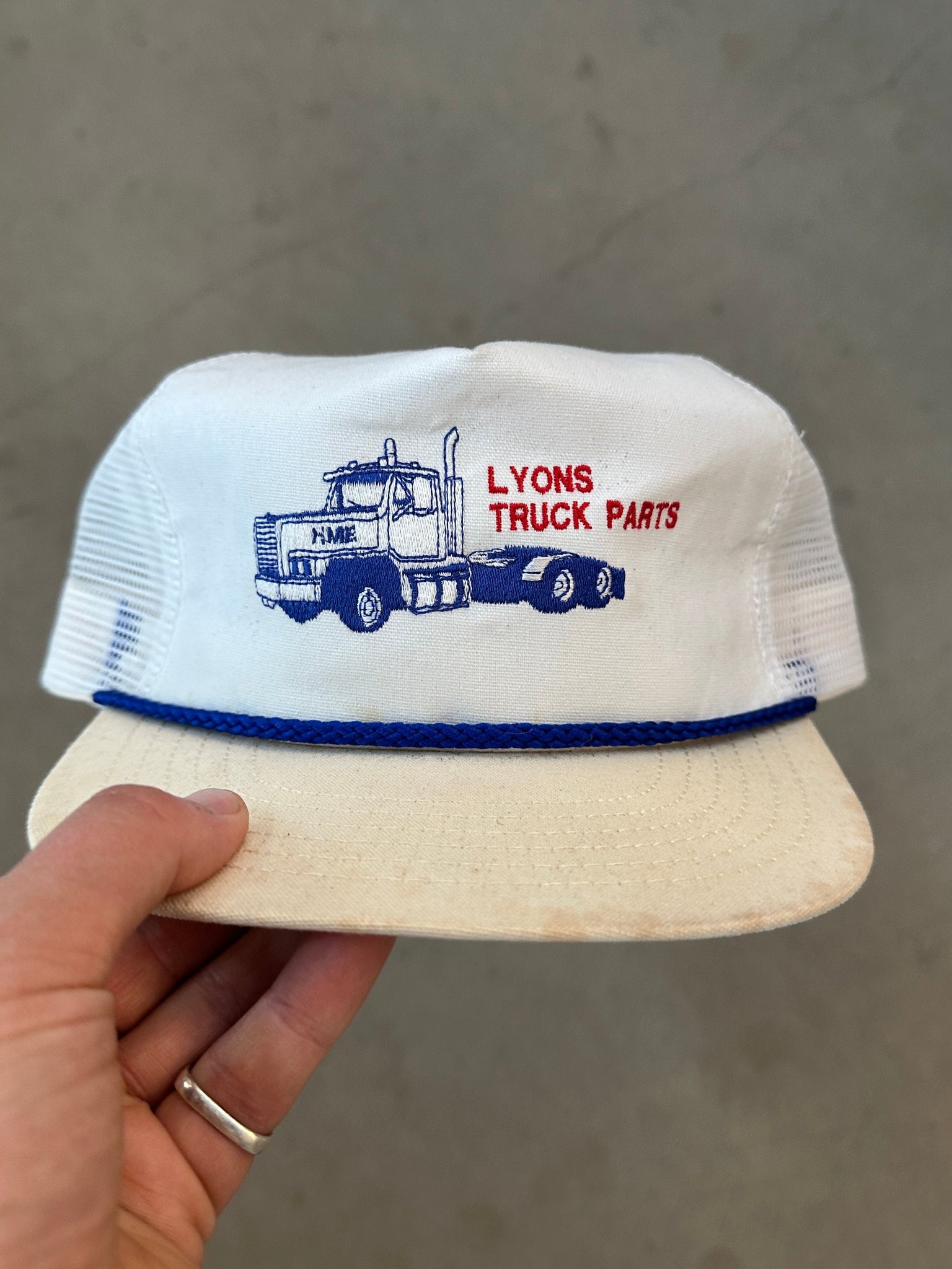 White cap with blue truck design and red text held by a hand against a gray background