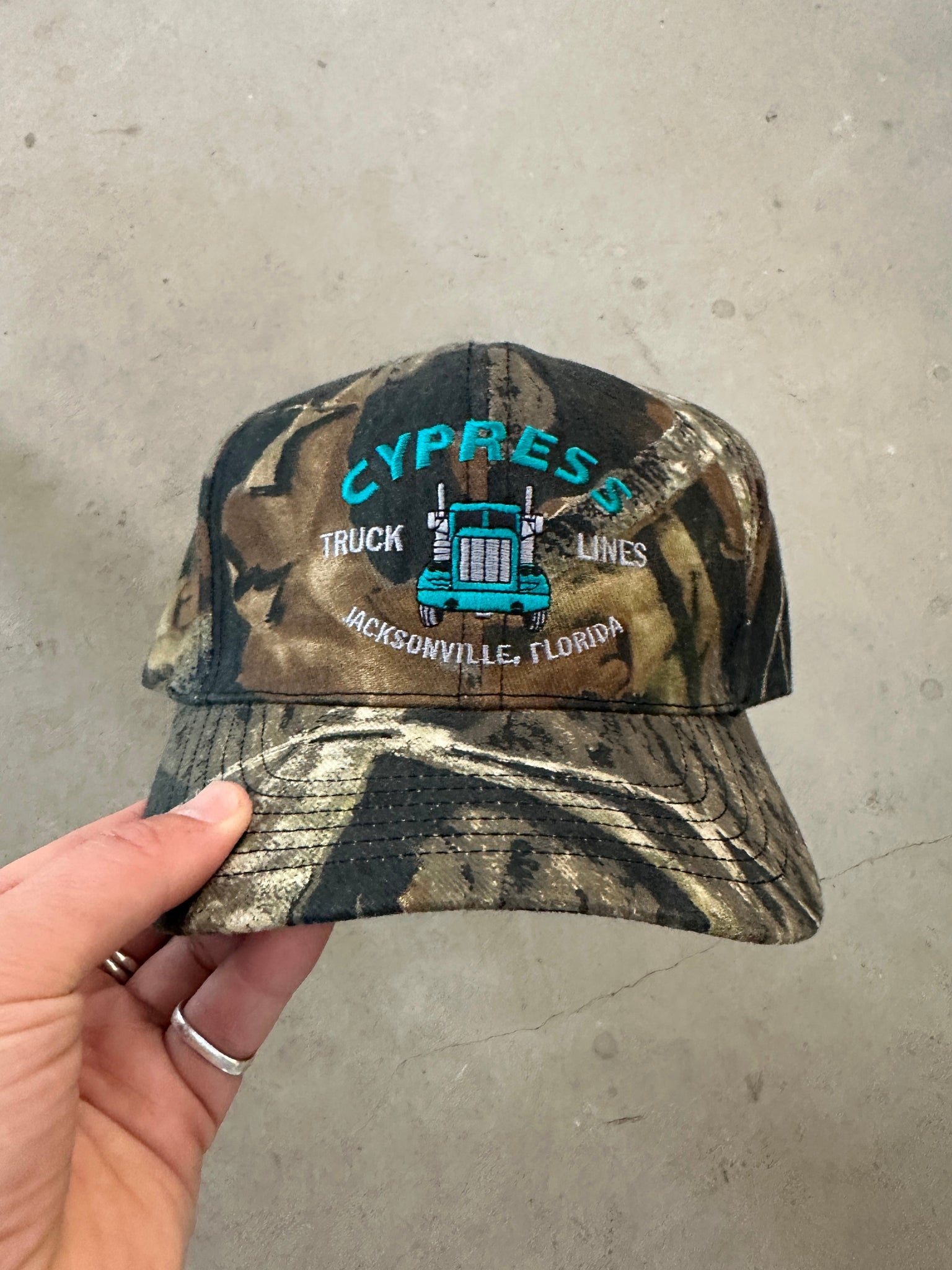 Camouflage cap with embroidered text held by a hand on a concrete floor.