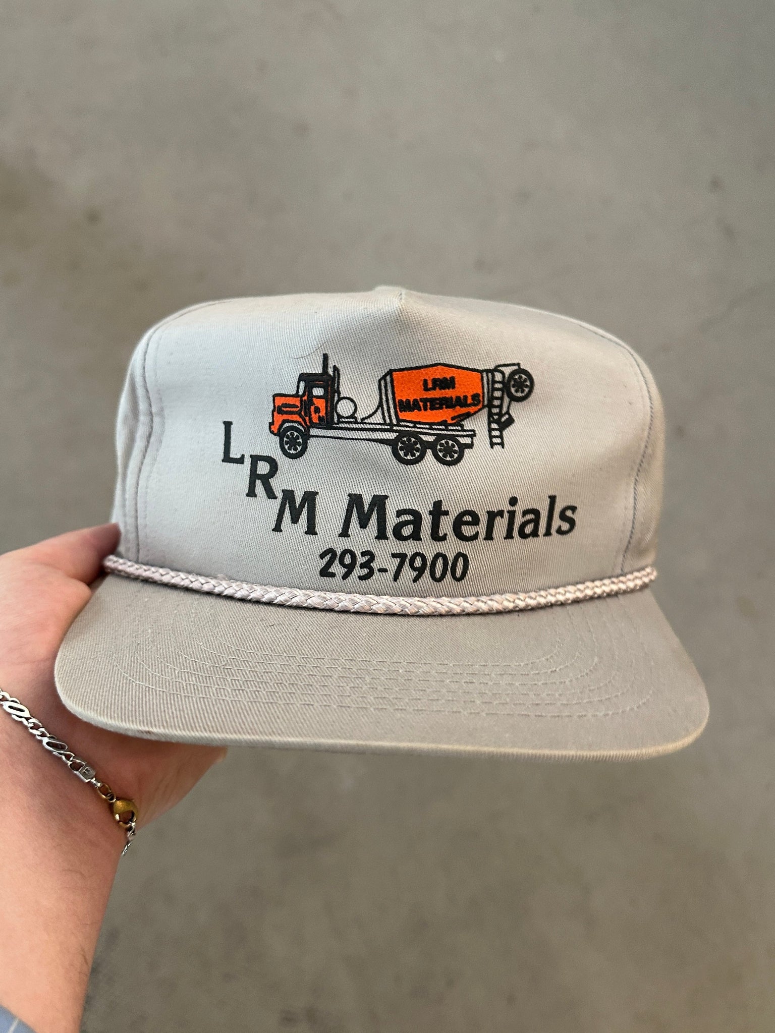 Gray cap with LR M Materials logo and contact information held by a hand against a gray background