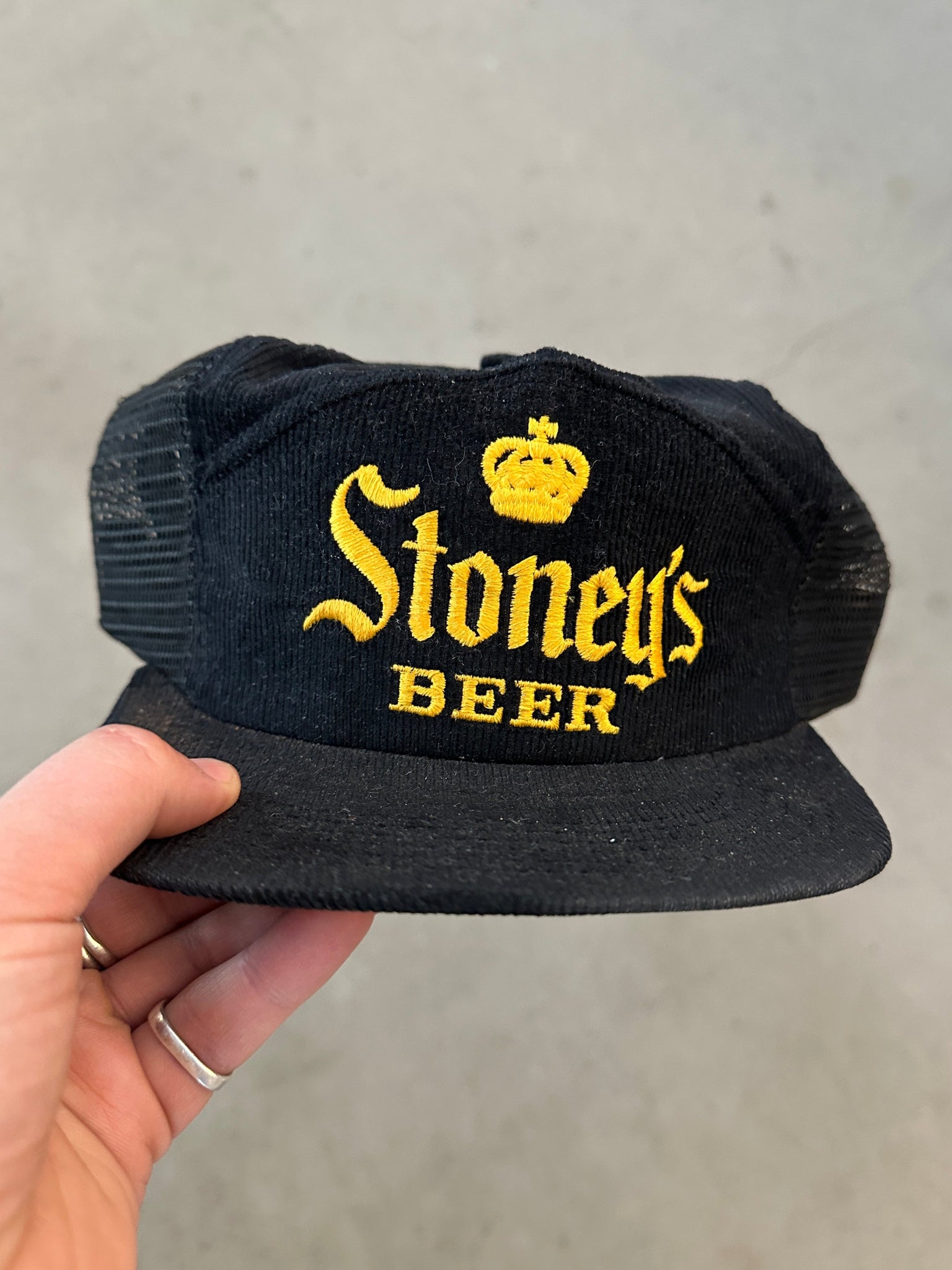 Black cap with 'Stoney's Beer' text held by a hand against a gray background