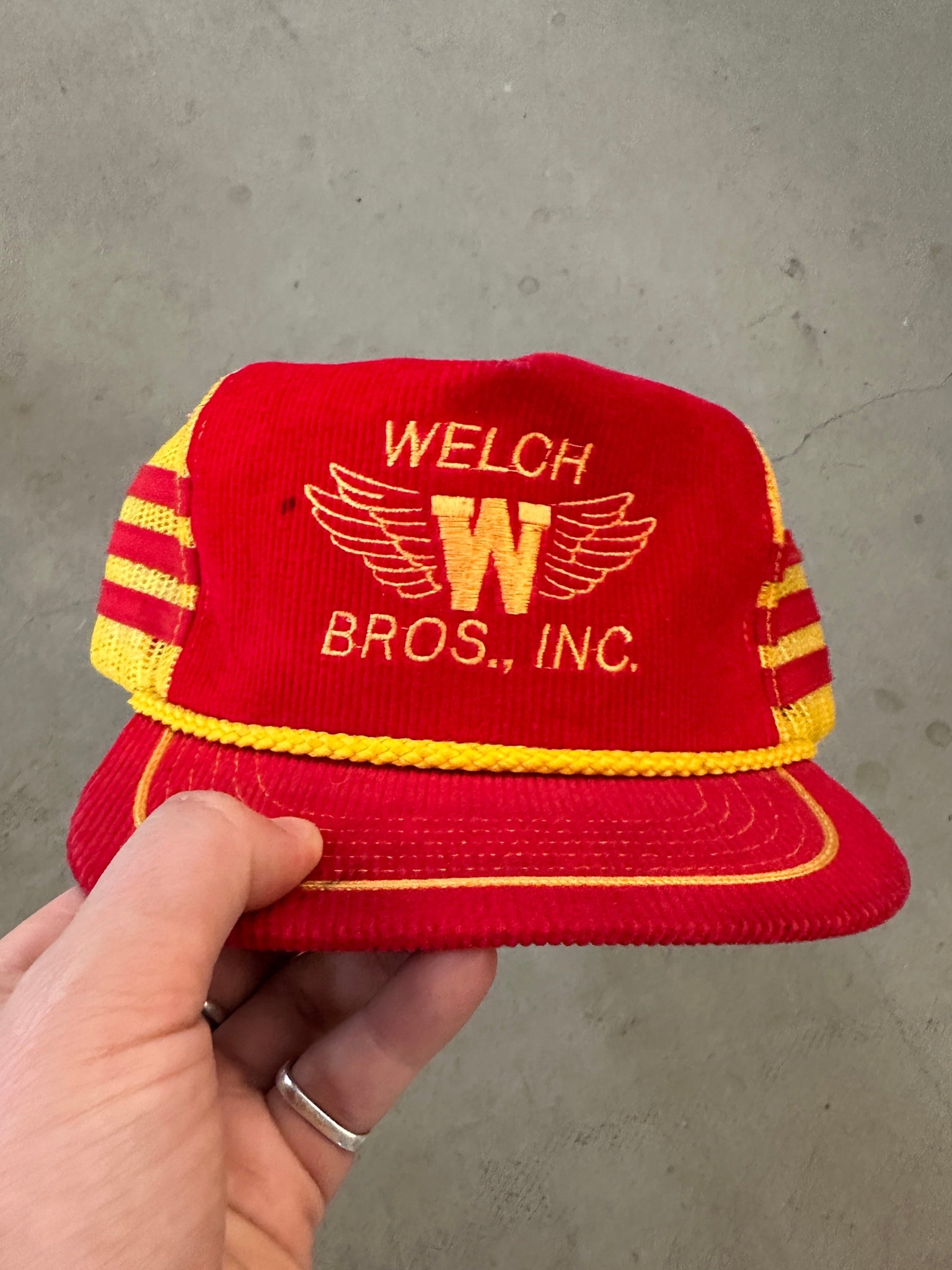 Red and yellow cap with 'Welch Bros., Inc.' logo held by a hand against a gray background