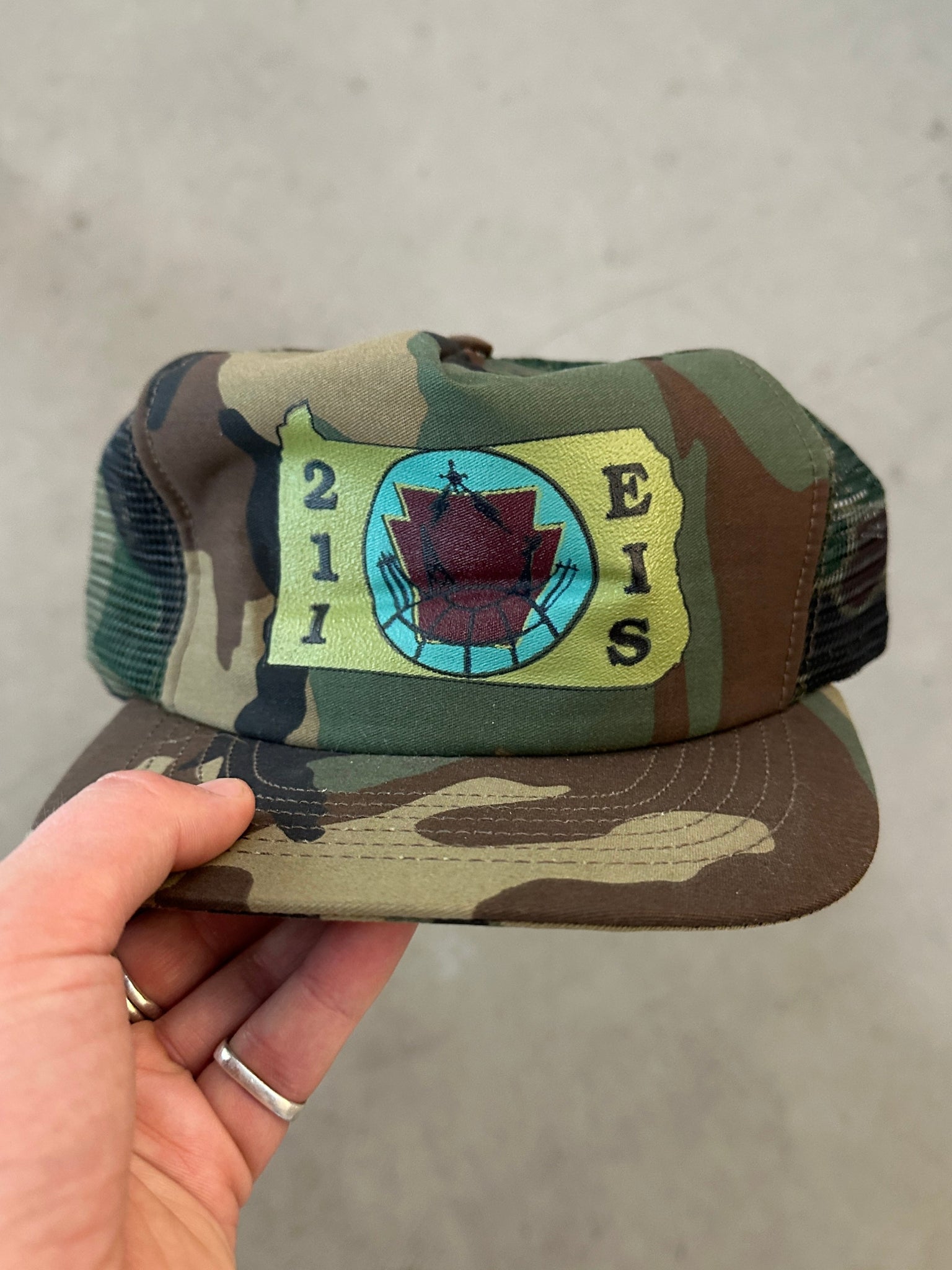 Camouflage cap with a logo and text held by a hand on a neutral background