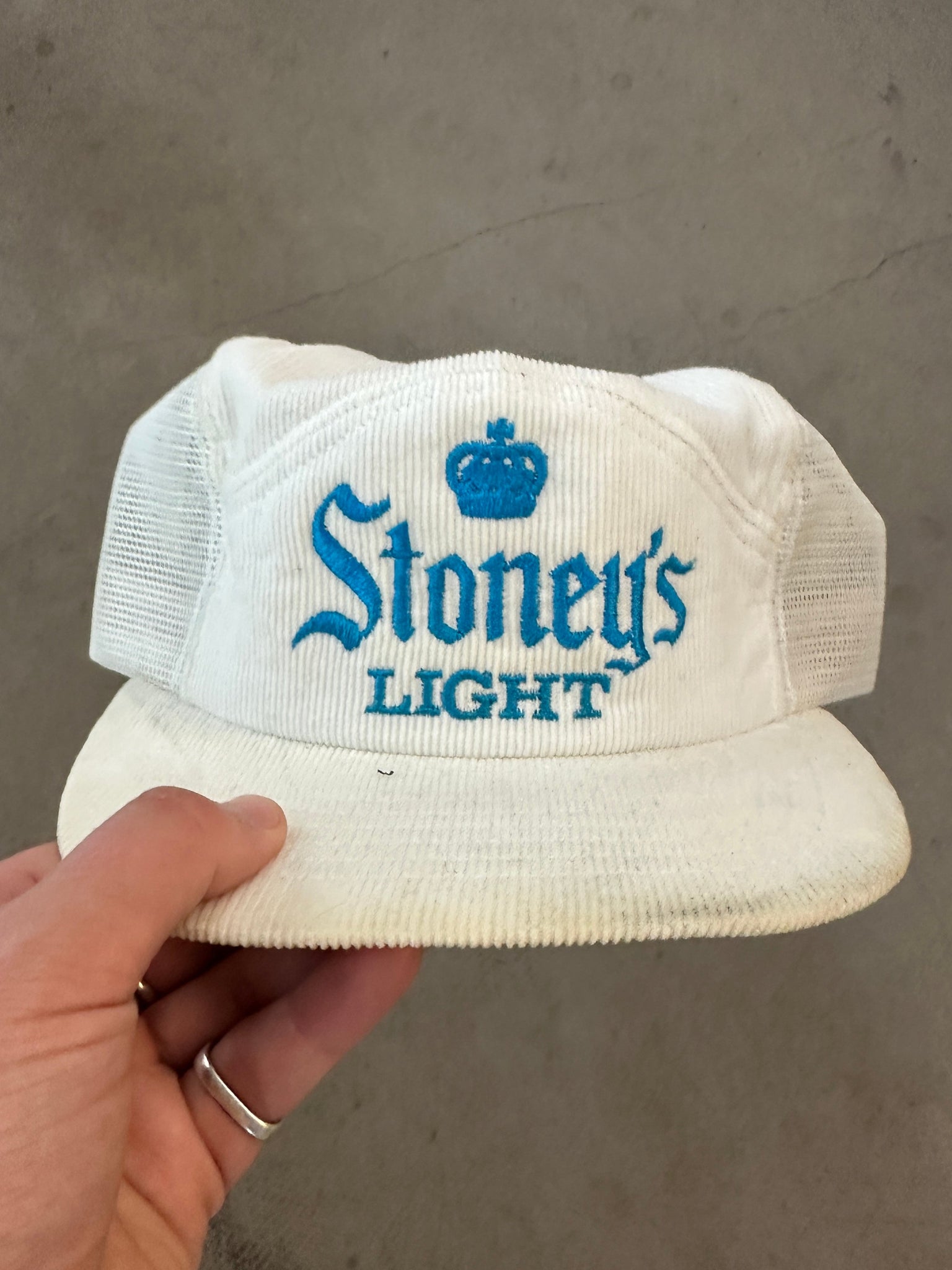 White cap with 'Stoney's Light' branding held by a hand against a gray background